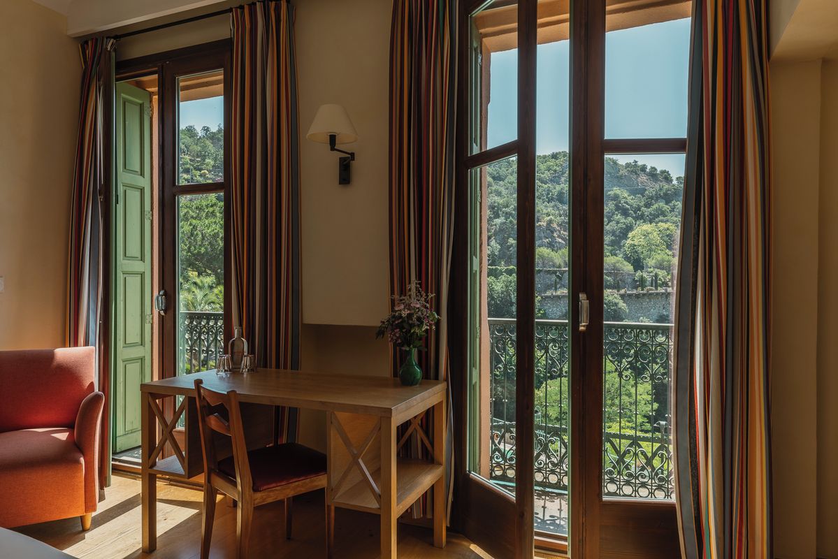 Een kamer met een houten tafel en stoelen, een groene deur en uitzicht op een weelderig landschap door grote ramen.