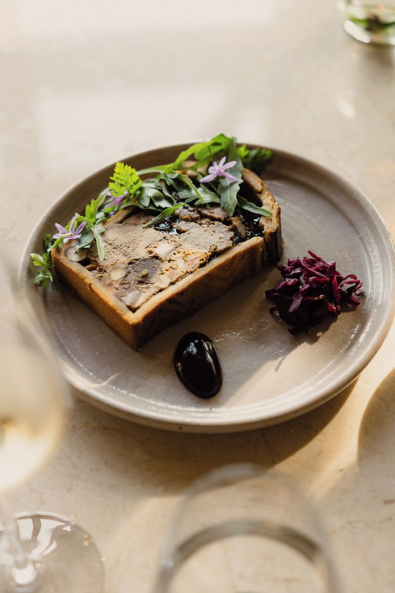A square-shaped food item with a crust, garnished with greens and purple flowers, on a round plate.