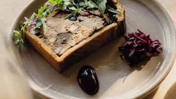 A square-shaped food item with a crust, garnished with greens and purple flowers, on a round plate.