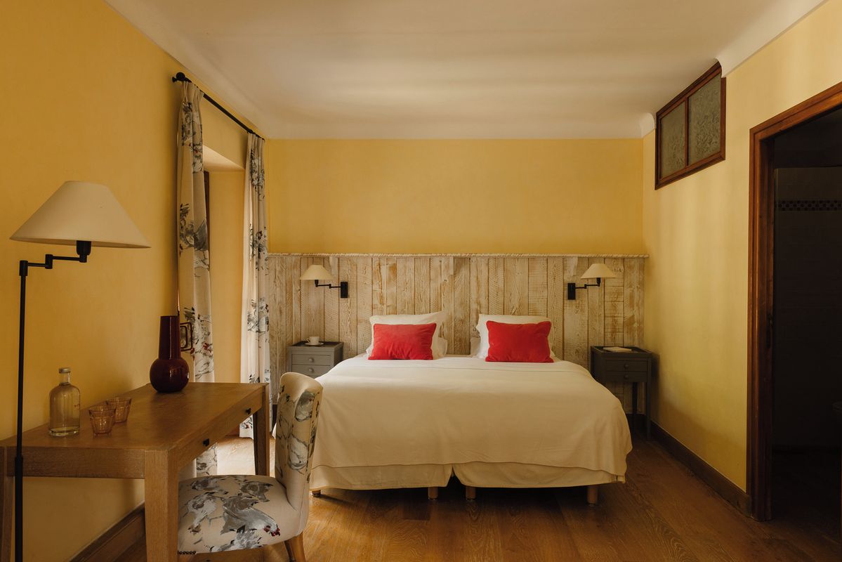 A bedroom with a wooden headboard, a bed with red pillows, a bedside table with a lamp, and a chair with a floral cushion.
