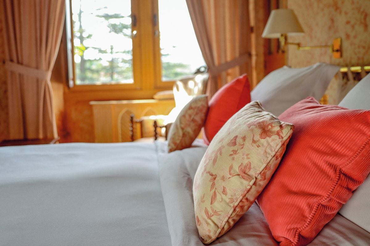 A neatly made bed with floral and solid pillows, a wooden headboard, and a window with curtains.