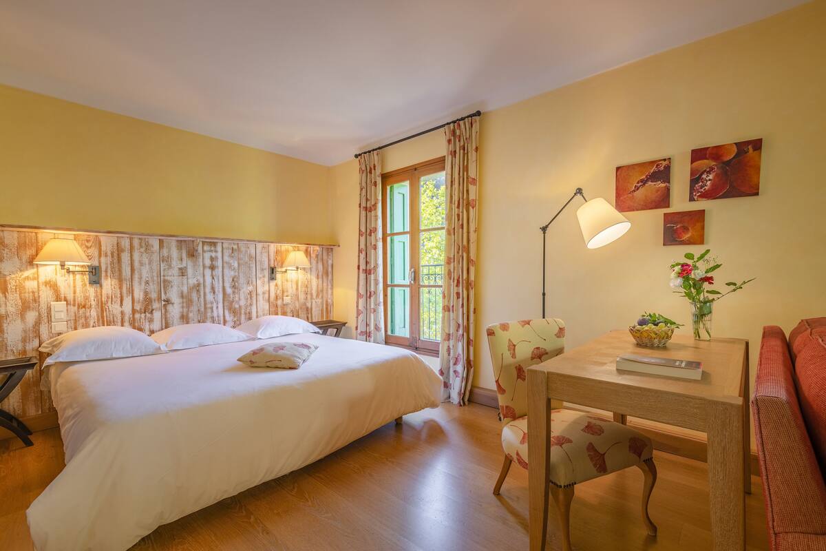Un dormitorio con cama, lámparas de noche, una ventana con cortinas, una mesa con un jarrón y una silla.
