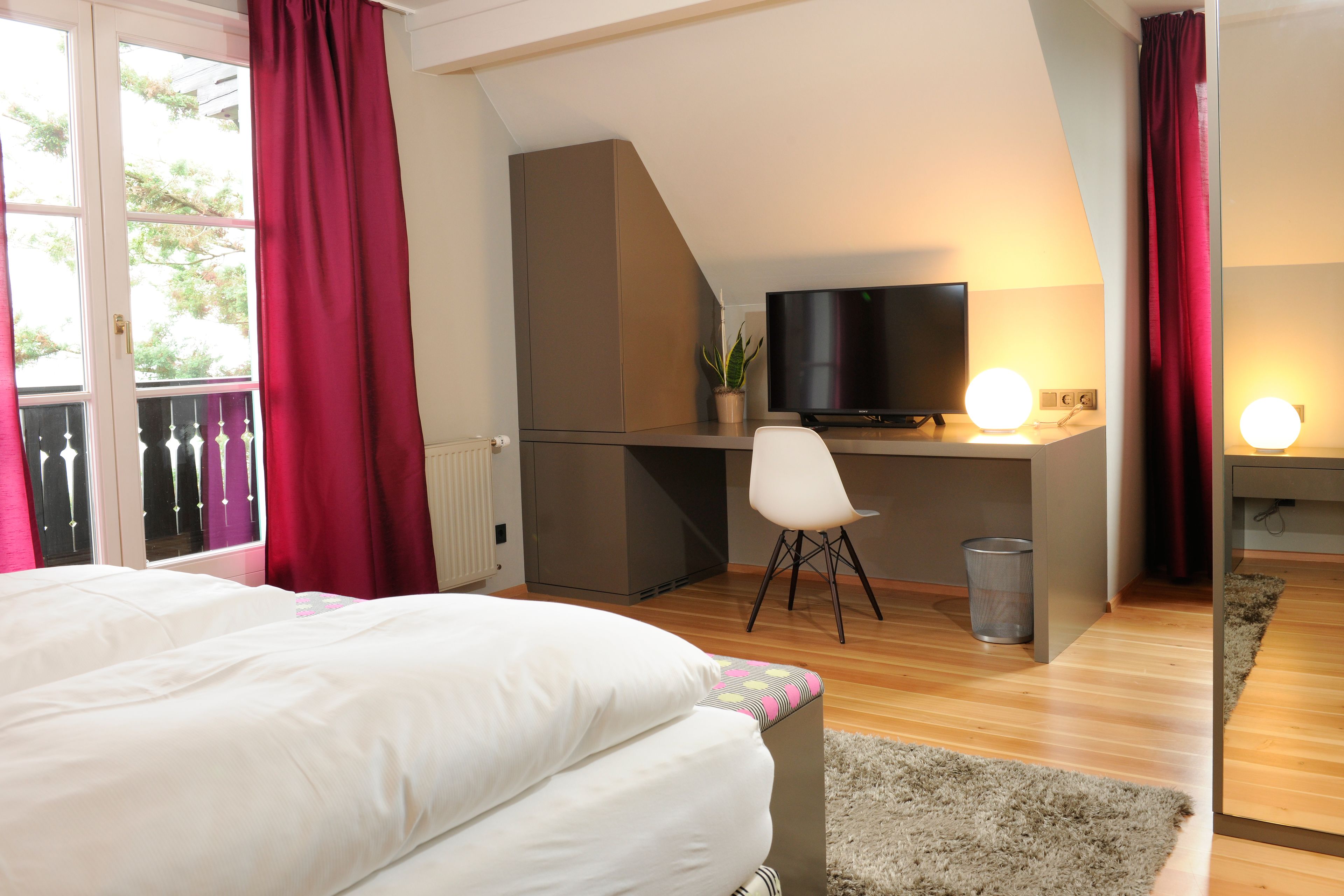 A bedroom with a bed, a desk with a chair, a television, and a window with curtains.