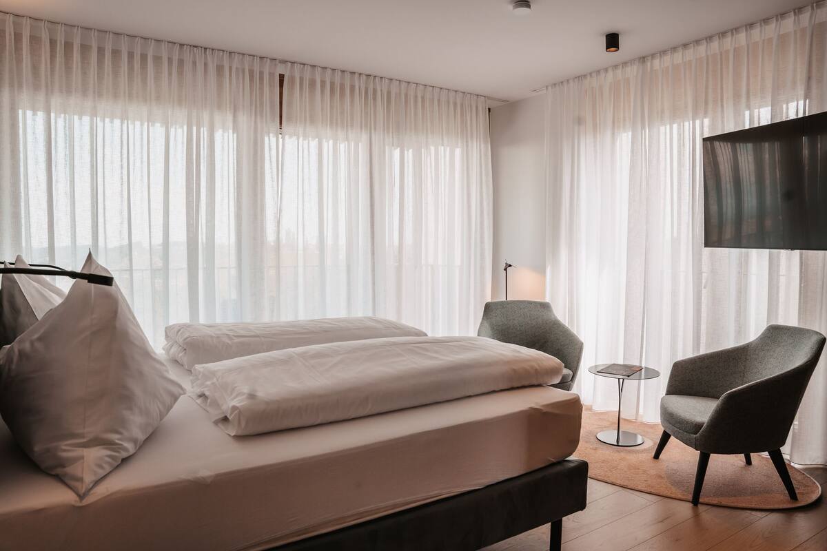 A hotel room with a bed, two armchairs, a small table, and a TV mounted on the wall.