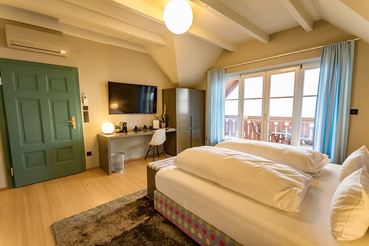 A bedroom with a bed, a desk, a chair, a TV, and a balcony with a railing.