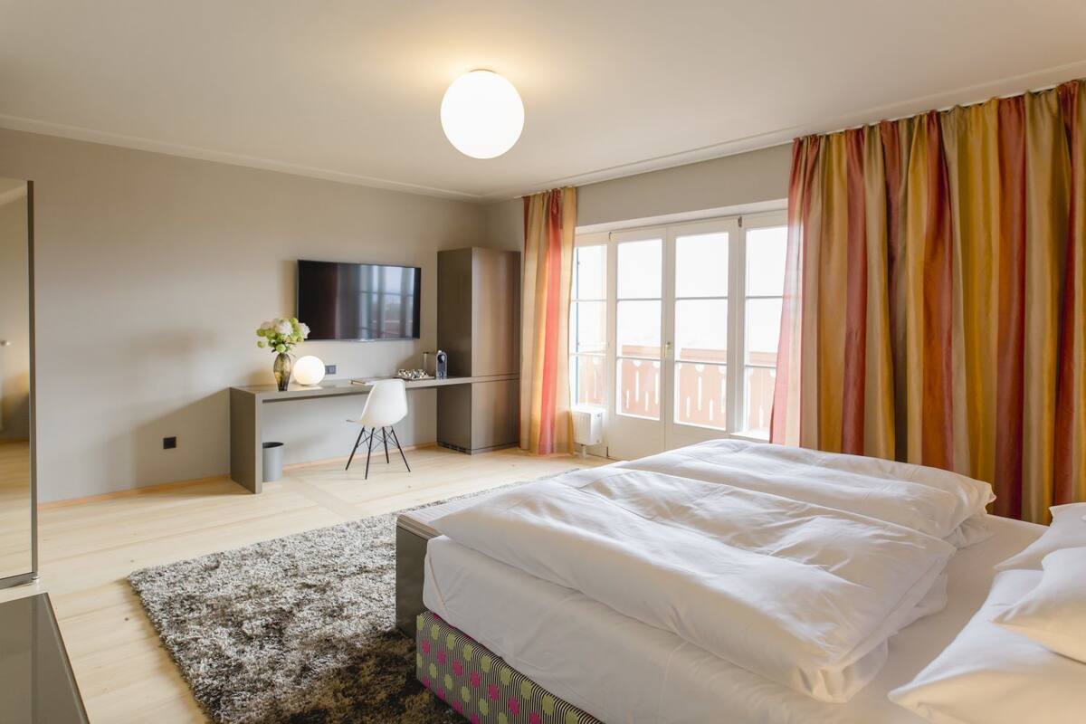 A modern bedroom with a large bed, a desk, a chair, and a TV.