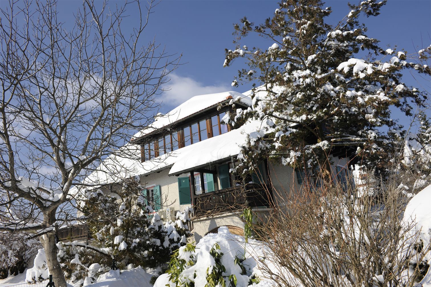 Ein mit Schnee bedecktes Haus mit Balkon, umgeben von Bäumen und Sträuchern.