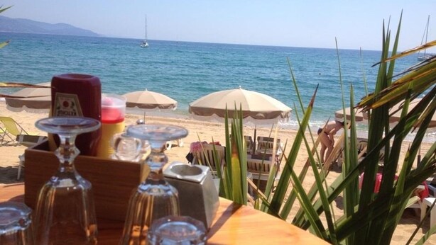 A beachside table with various glassware and a bottle, set against a backdrop of the sea and umbrellas.