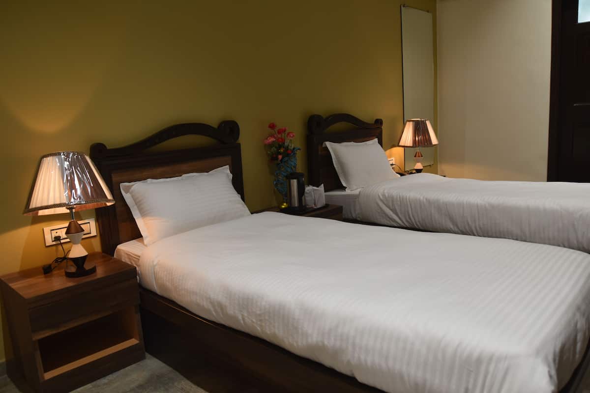 Uma cama de casal com roupa de cama branca, uma mesa de cabeceira com abajur e um vaso com flores.