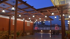 Un espacio patio al aire libre con piso de madera, una pérgola con luces colgantes y vista a un edificio con cúpula al fondo.