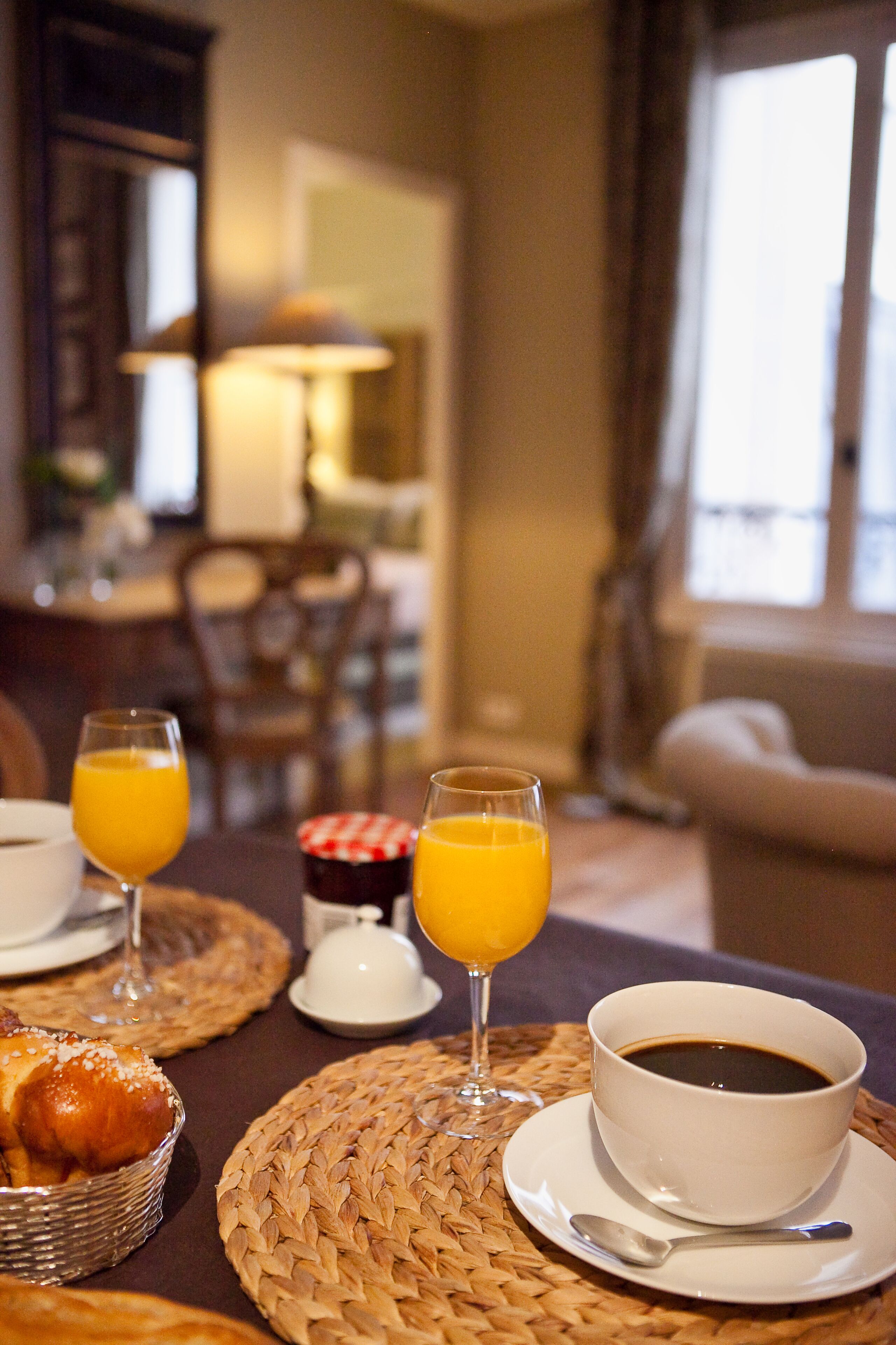 A breakfast setting with a coffee cup, two glasses of orange juice, and a basket of pastries on a woven placemat.