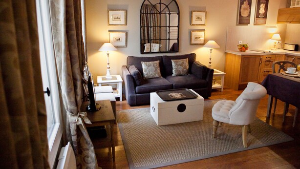 Un salon avec un canapé de couleur foncée, une table d’appoint blanche, une table basse et un fauteuil. Un grand miroir est accroché au mur, et des tableaux encadrés sont disposés au-dessus du canapé.