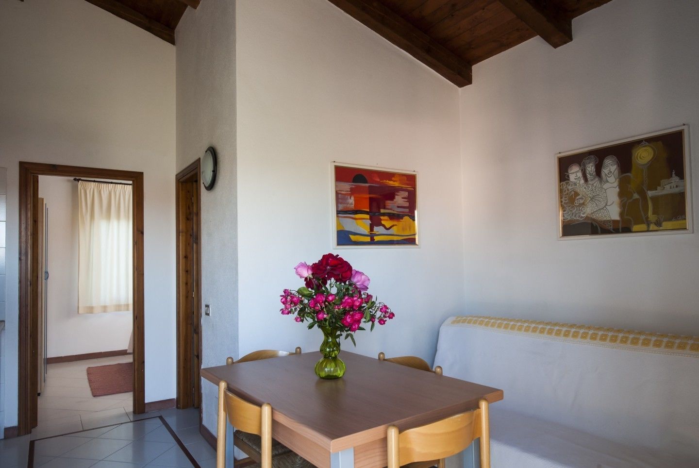 A dining area with a wooden table, chairs, a vase of flowers, a painting, and a sofa.