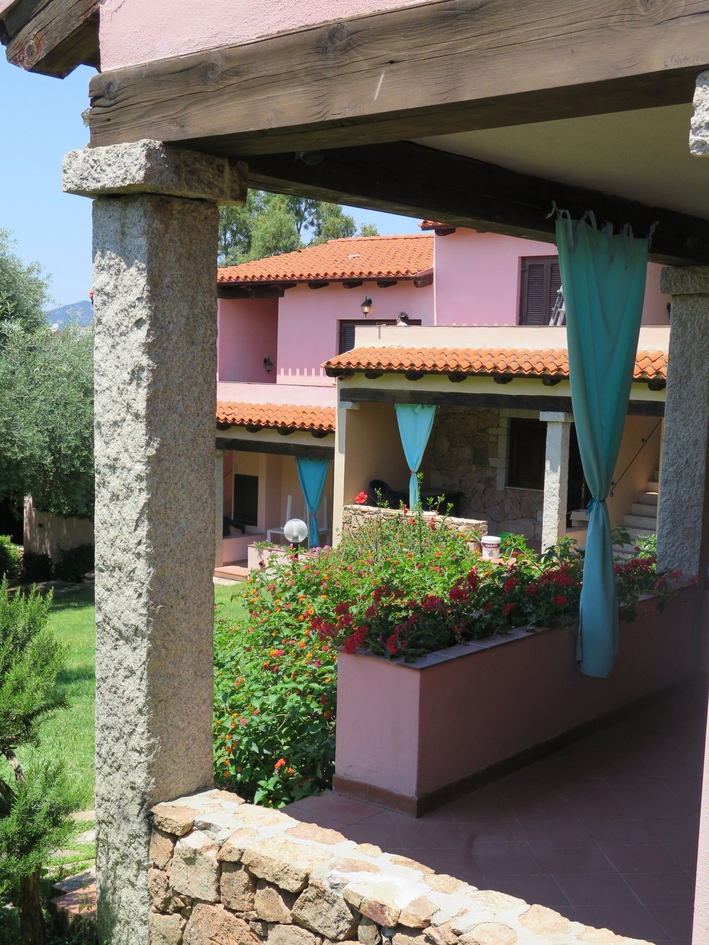A covered outdoor area with a stone wall, a planter with flowers, and a building with a tiled roof.