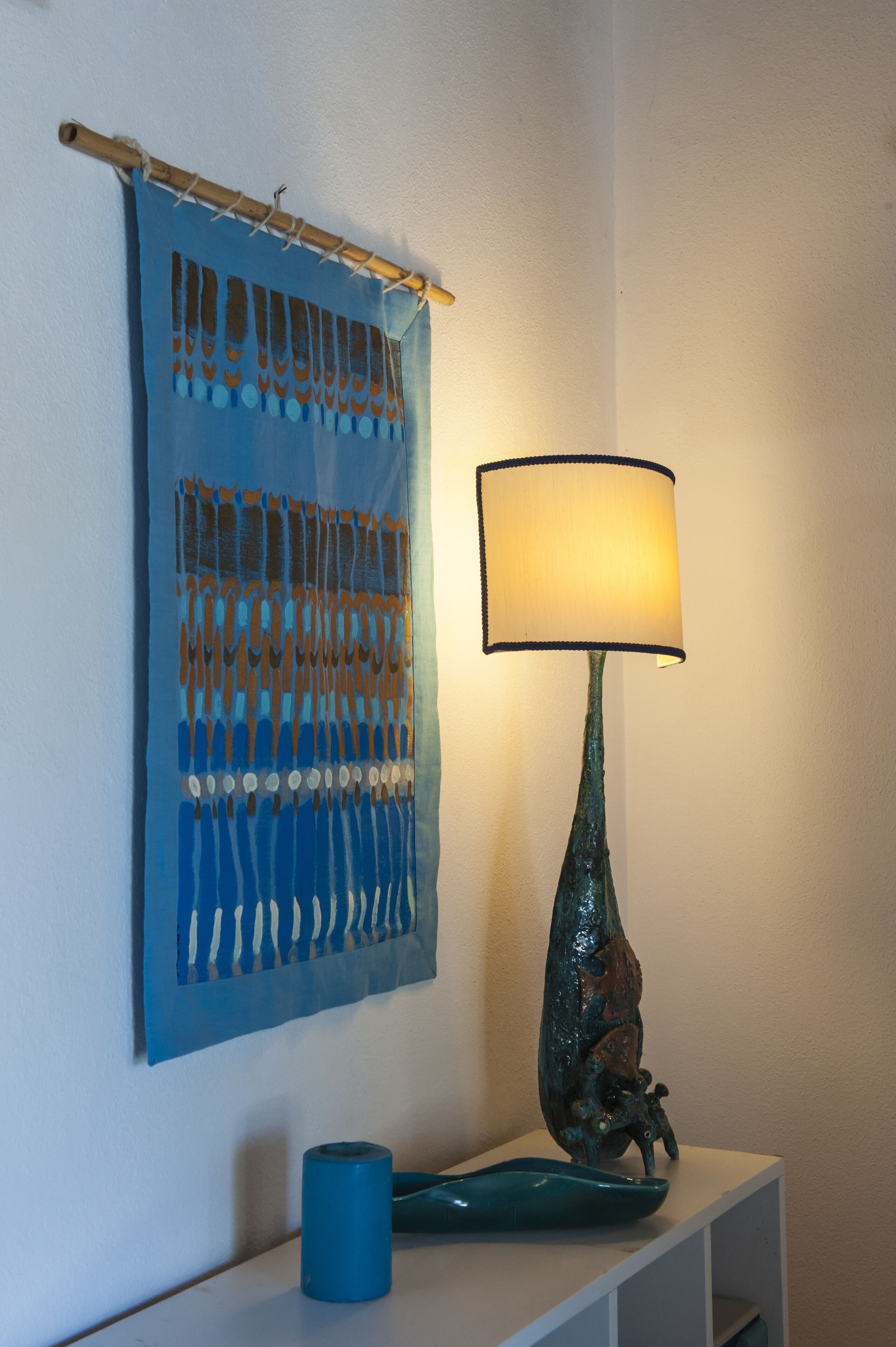 Une tapisserie murale ornée d’un motif géométrique, une lampe de table avec un pied bleu et une figurine de grenouille décorative, ainsi qu’un objet cylindrique bleu posé sur une étagère blanche.