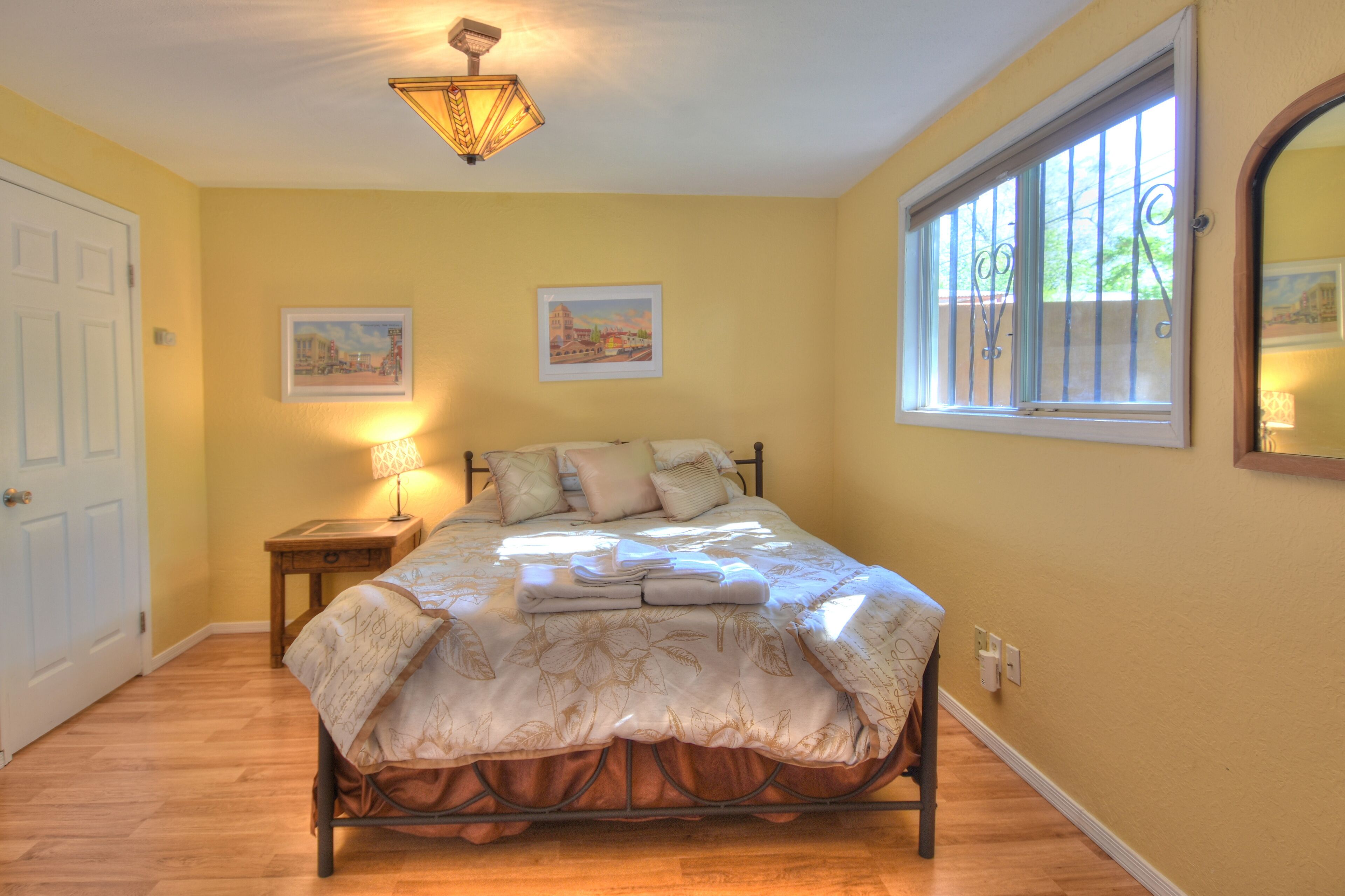 Un dormitorio con cama, mesita de noche, lámpara, ventana con barrotes y dos cuadros en la pared.