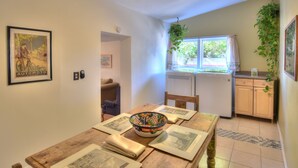 Ein Essbereich mit einem Holztisch, Stühlen und einer Schüssel. Ein Fenster mit Vorhängen, ein Deckenventilator und eine Küchenzeile mit Schränken und einem Kühlschrank.