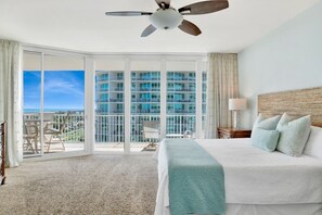 A hotel room with a bed, a ceiling fan, a view of a high-rise building, and a balcony with chairs.