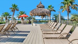 Una terraza de madera con sillones de descanso y una estructura con techo de paja, con vistas a una playa con palmeras y un cielo azul despejado.