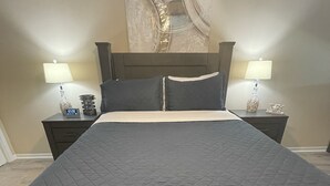 Una cama bien hecha con un edredón oscuro, dos mesitas de noche con lámparas, un reloj y un jarrón decorativo.