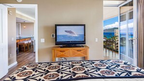 Habitación de hotel con cama, un televisor mostrando una ballena, balcón con vista al mar y un comedor visible a través de una puerta abierta.
