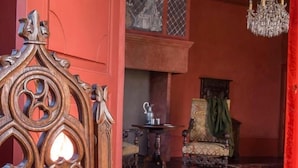 Una habitación con pared roja, una silla de madera, una mesa con un libro abierto y gafas, y una araña.