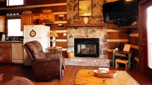 A rustic cabin interior with a stone fireplace, wooden walls, a leather chair, a wooden table, and a television mounted above the fireplace. - SERENITY IN THE MOUNTAINS - Private-view-convenient-unique-hot tub-ev charging (Sevierville)