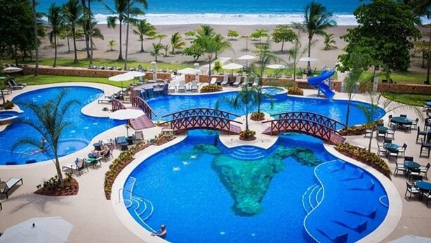 A large, multi-level swimming pool with a bridge connecting different sections, surrounded by lounge chairs and umbrellas. - Beachfront Oasis~ Huge Terrace ~ Amazing Sunsets~ Resort Pool~ Grill~ Beach gear (Jacó)