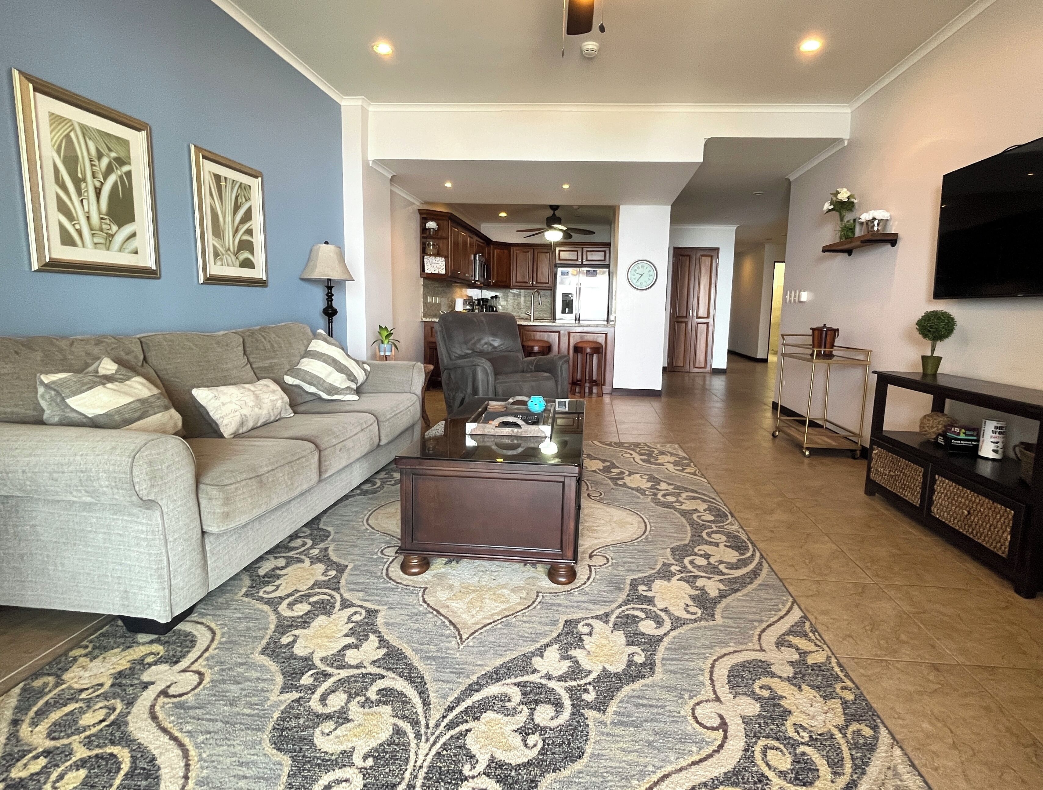 A living room with a sofa, coffee table, and rug. There is a kitchen in the background with cabinets and a clock.