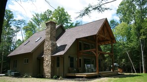 A two-story log cabin with a stone chimney, a porch with outdoor seating, and a large window overlooking the surrounding forest.