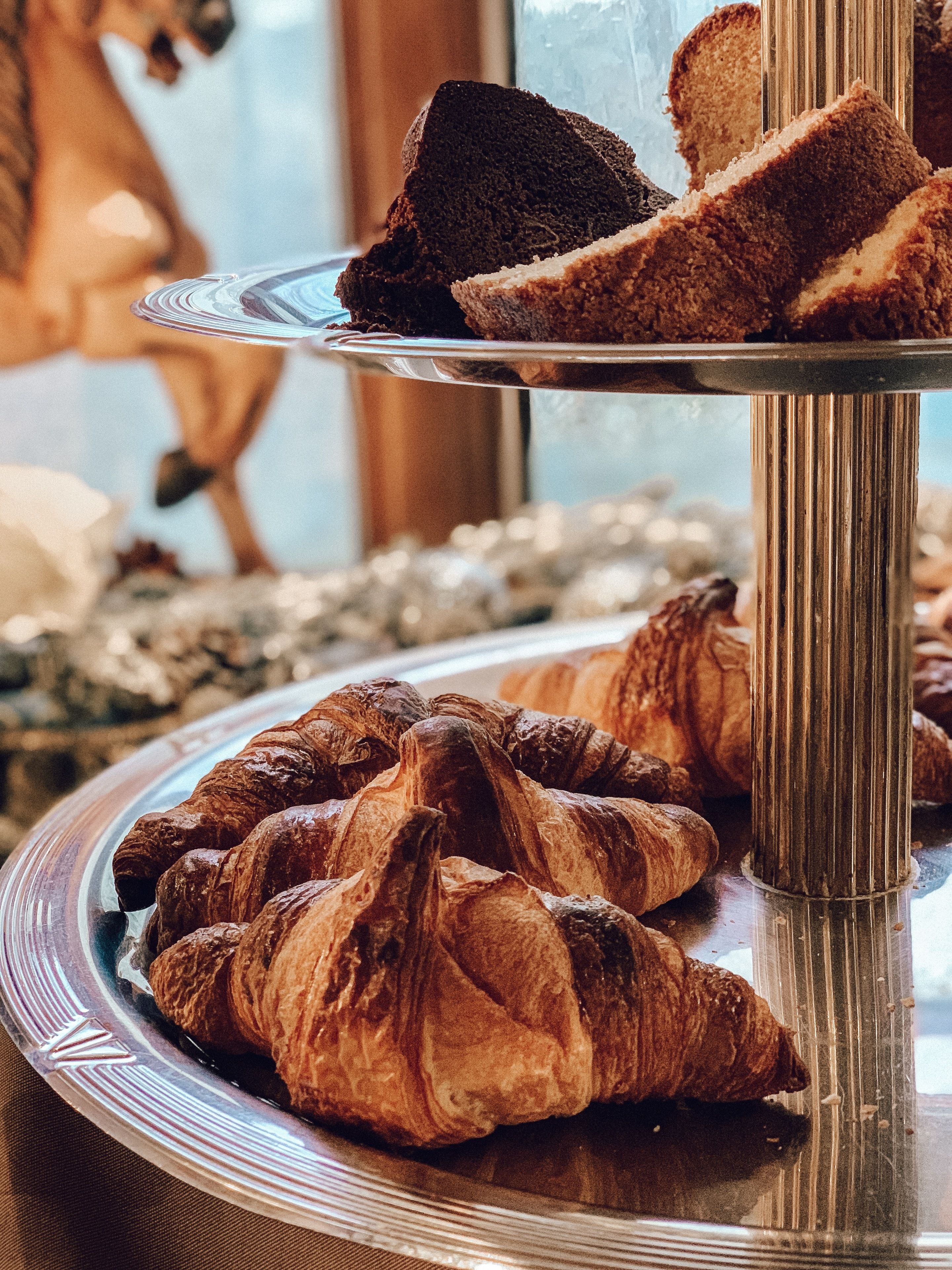 A display of pastries, including croissants and slices of cake, on a reflective metal stand.