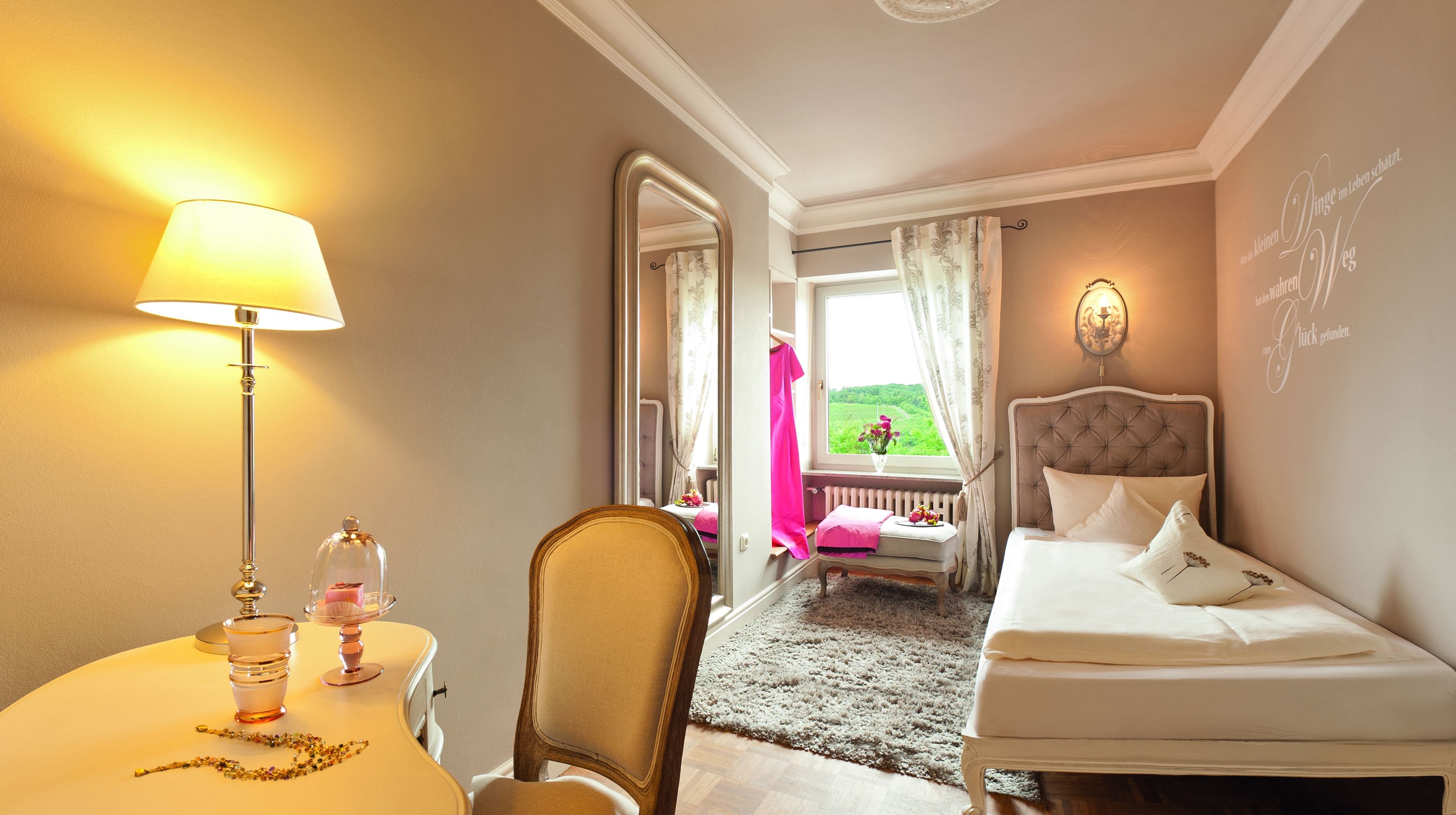 Habitación de hotel con cama, una silla, una mesa con lámpara y vistas a un jardín por la ventana.