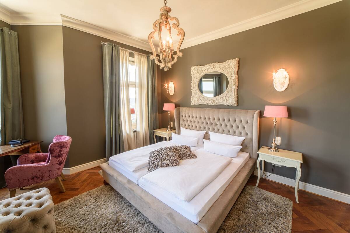 Une chambre à coucher avec un grand lit, une tête de lit capitonnée, deux tables de chevet avec des lampes, un miroir au mur et une chaise.