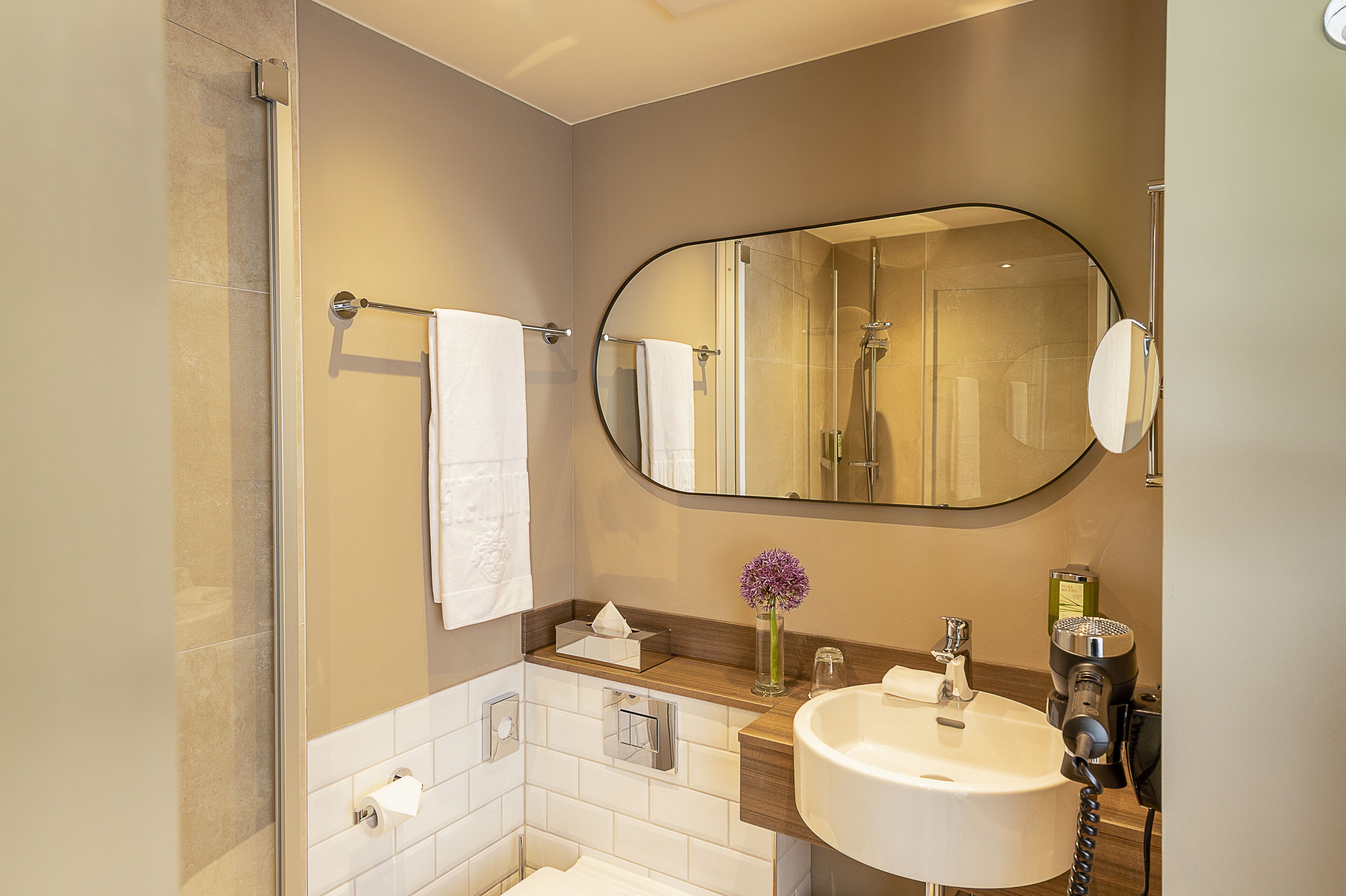 A modern bathroom with a glass shower enclosure, a large oval mirror, a white sink, and a wooden vanity.
