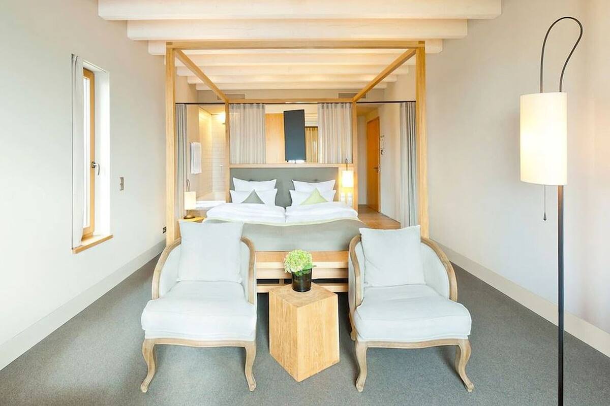 Une chambre d’hôtel avec un grand lit, deux fauteuils, une petite table en bois et une lampe sur pied.