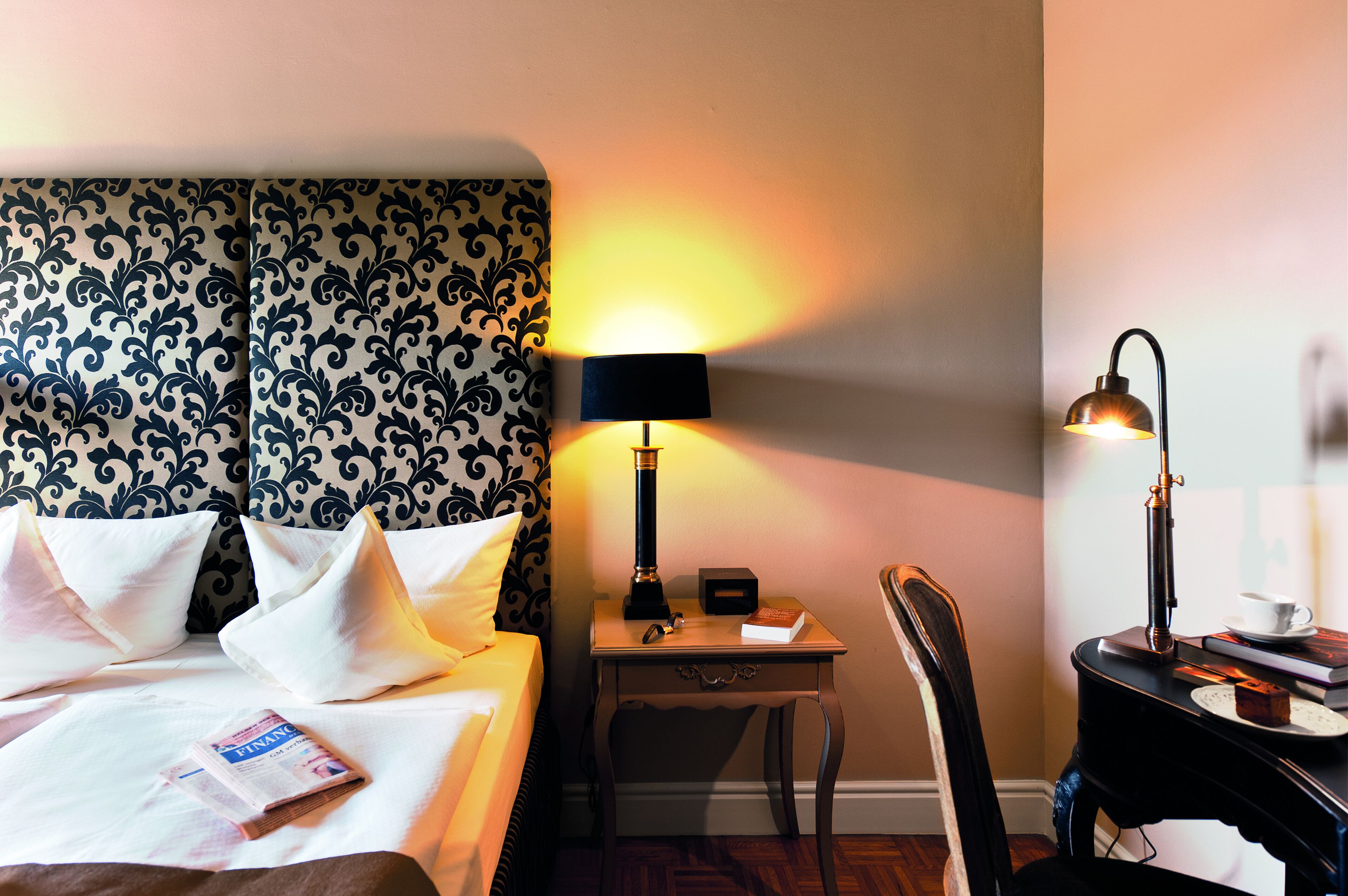 A hotel room with a bed, a nightstand with a lamp, a chair, and a small table with a cup and saucer.
