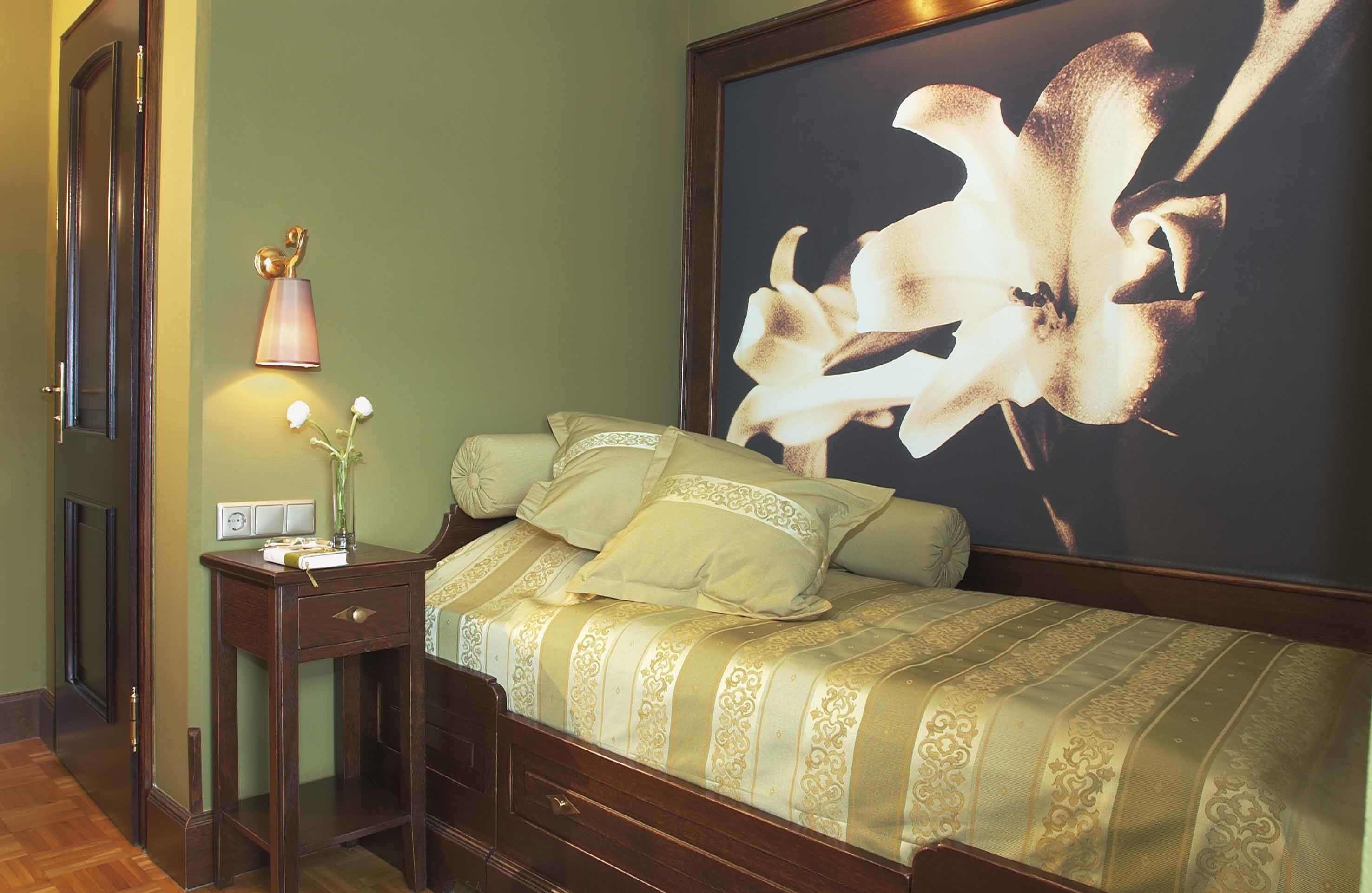 Un letto rifatto con cura, con una coperta a fiori, un comodino con una lampada e un grande quadro floreale appeso al muro.