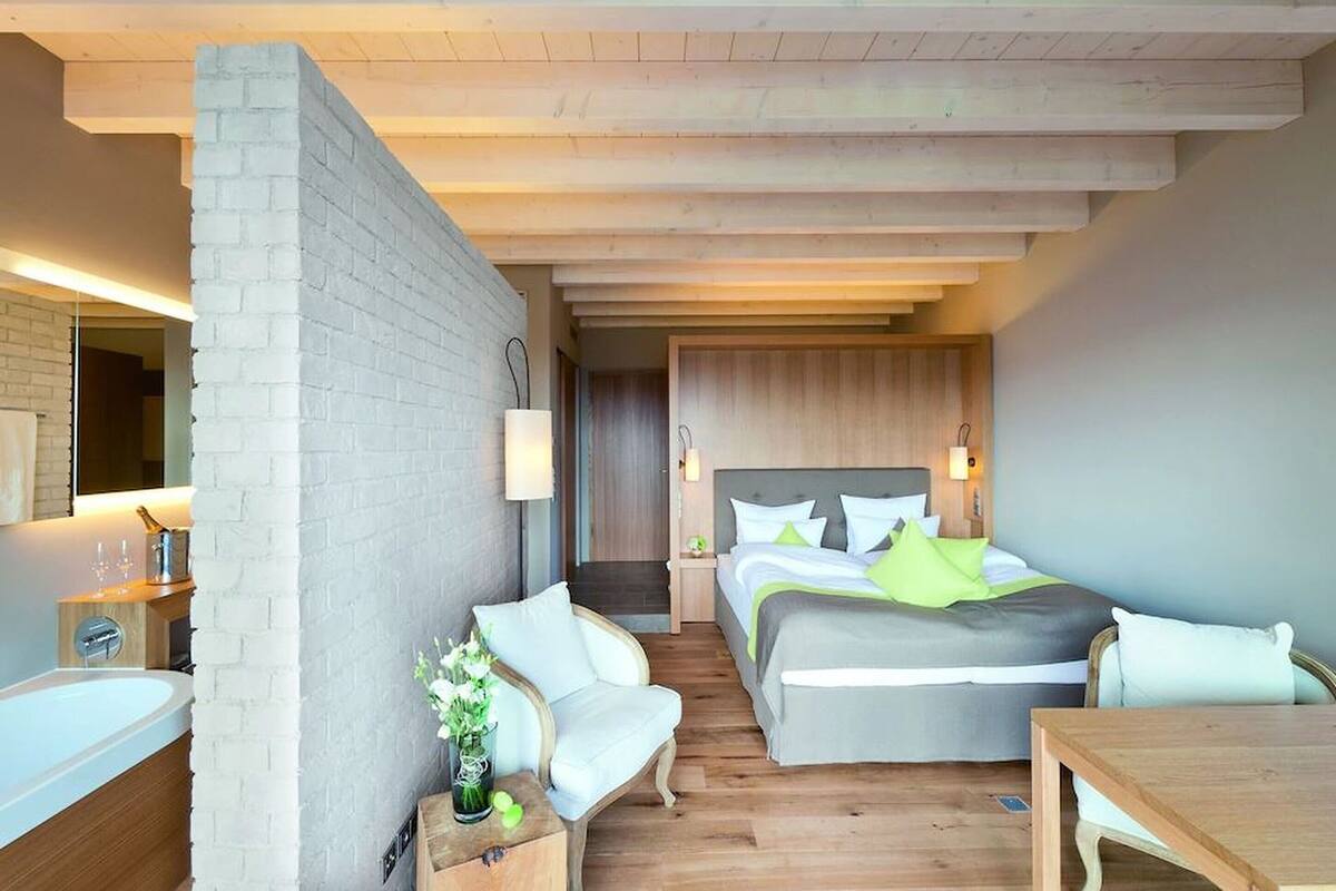 Une chambre d’hôtel moderne dotée d’un grand lit, d’un coin salon avec un fauteuil, une petite table et d’une salle de bain avec baignoire.