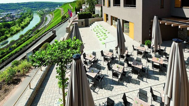 An outdoor terrace with tables and chairs, overlooking a river and greenery.