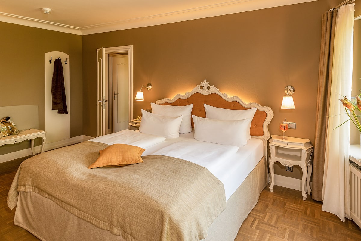 Un lit double avec du linge de lit blanc, deux tables de chevet avec des lampes, une tête de lit en bois et une vue sur une fenêtre avec des rideaux.