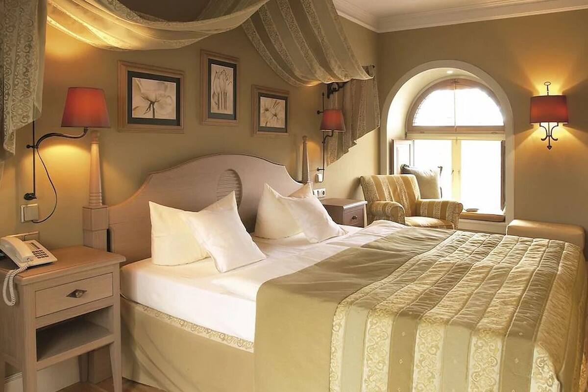 Une chambre d’hôtel avec un grand lit, une table de chevet avec une lampe, une chaise et une fenêtre avec des rideaux.