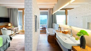 A modern hotel room with a large window, a bathtub, a vanity with a mirror, and a seating area with a TV and a small table.