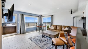 A spacious living area with a large window, a brown leather sofa, a wooden coffee table, and a dining table with chairs.