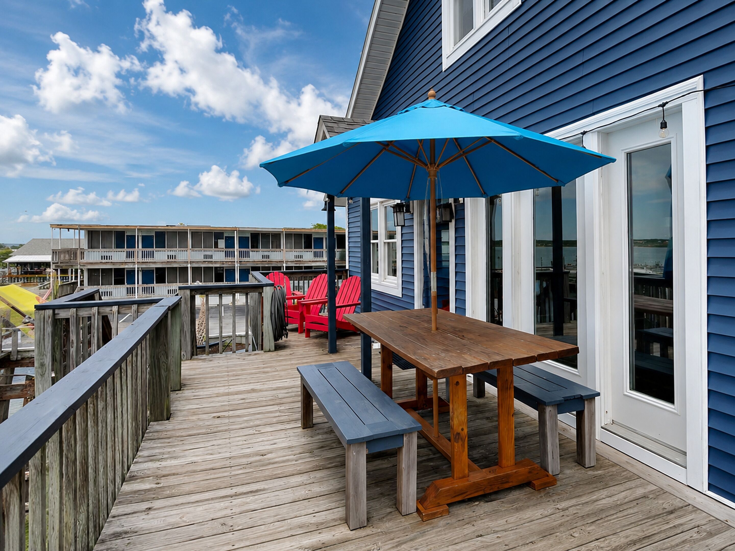 A wooden deck with a table and benches, a blue umbrella, and a building with multiple windows in the background.