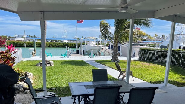 Un porche cubierto con una mesa y sillas, con vista a un área de piscina con bandera, palmeras y barcos.