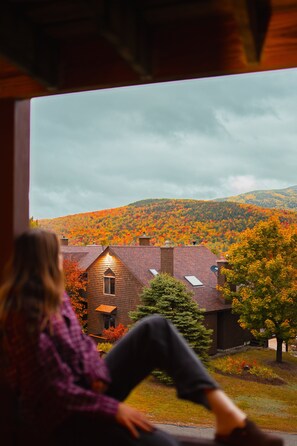 A person sitting by a window overlooking a house with a sloped roof and a view of a hillside with autumn foliage.