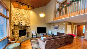 A spacious living room with a stone fireplace, wooden flooring, a large sectional sofa, and a balcony with festive decorations. - V3 Gorgeous Ski-In slopeside Townhouse with Amazing Mountain Views in Bretton Woods. Dog Friendly! (Bretton Woods)