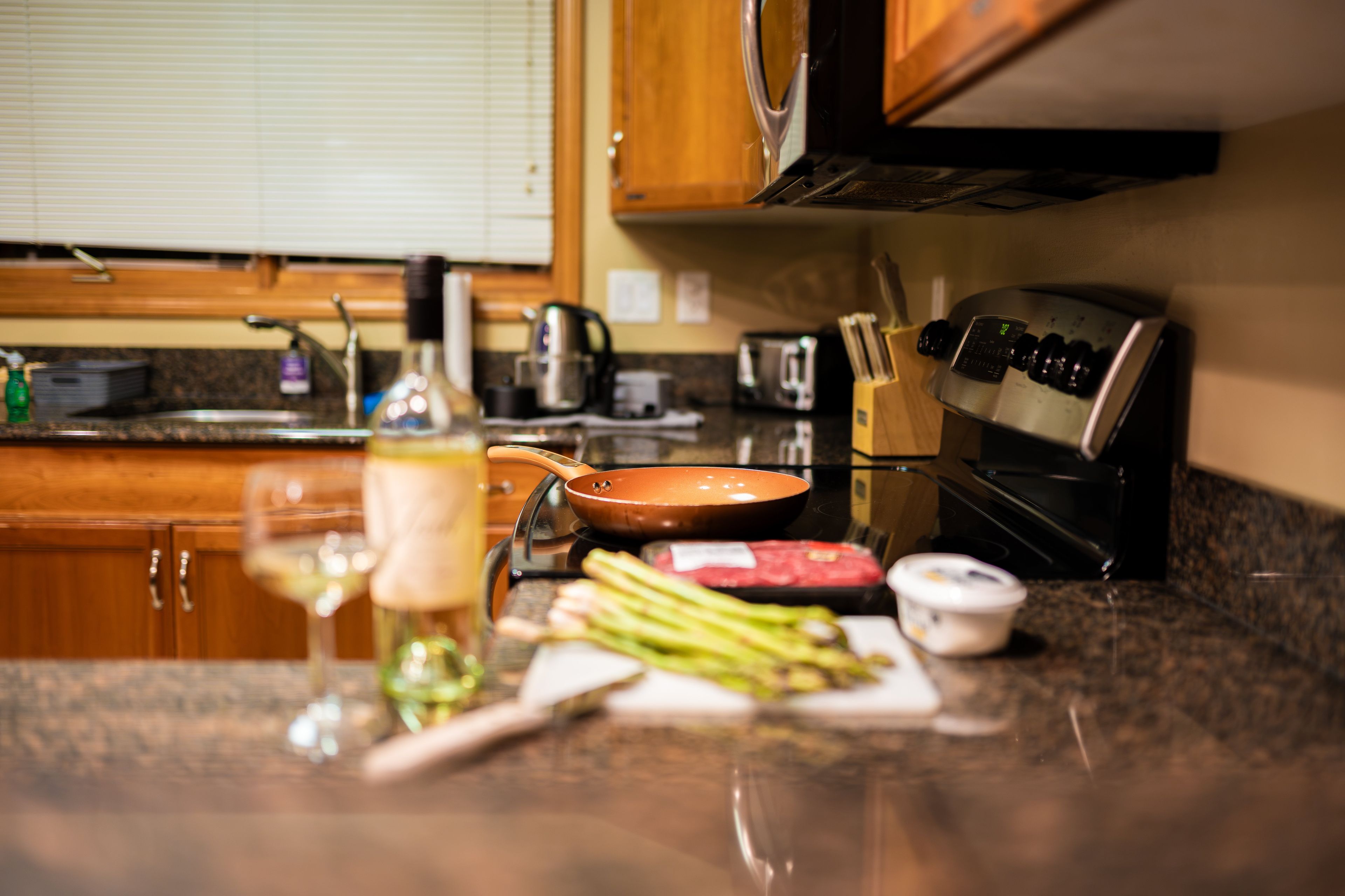A kitchen counter with a frying pan, a cutting board with meat, a bowl of vegetables, a glass of wine, and a coffee maker.