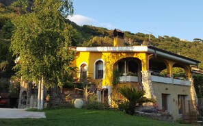 Una villa amarilla con ventanas arqueadas y fachada de piedra, rodeada de exuberante vegetación y una ladera.
