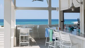 A covered outdoor seating area with white chairs and tables, a bar with various bottles, and a view of the ocean. - Last minute discount July 5-11!  Watercolor Camp district 5BR w/ Pool & Spa (Santa Rosa Beach)